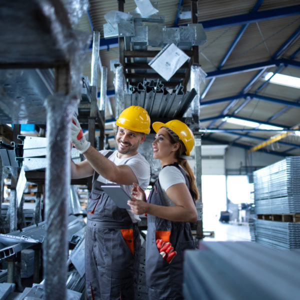 A man and woman in overalls among steel frames, counting stock items, and inputting it onto a tablet